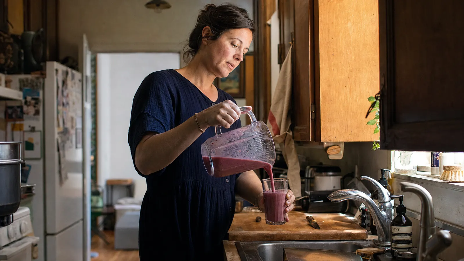 Woman pouring a finished smoothie in a narrow galley kitchen in morning light